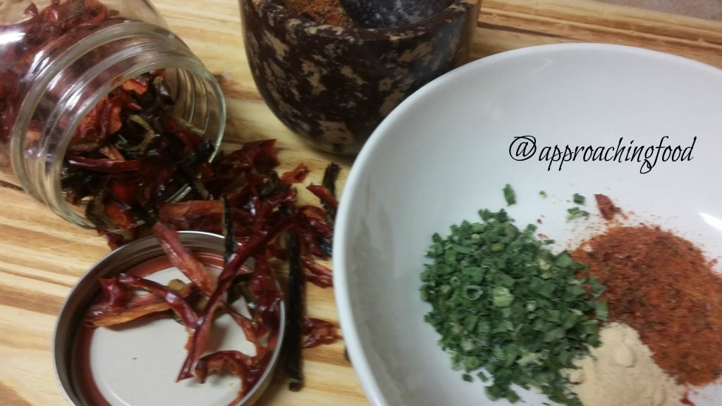 Dried chives, ground dehydrated peppers, and garlic powder are in a bowl. Dried peppers are spilling out of a mason jar on its side. 